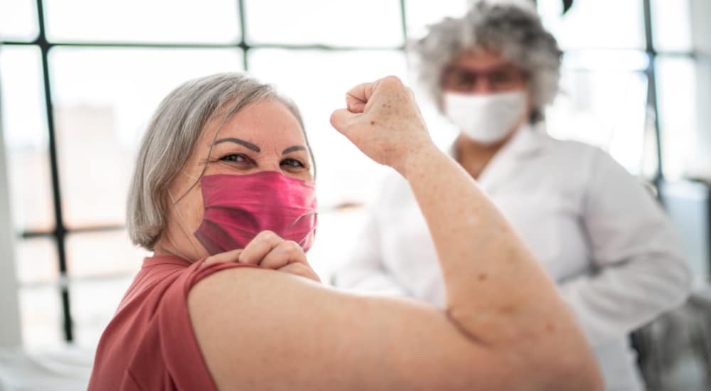 Une femme souriante et masquée montre son biceps, poing levé. En arrière plan une soignante.